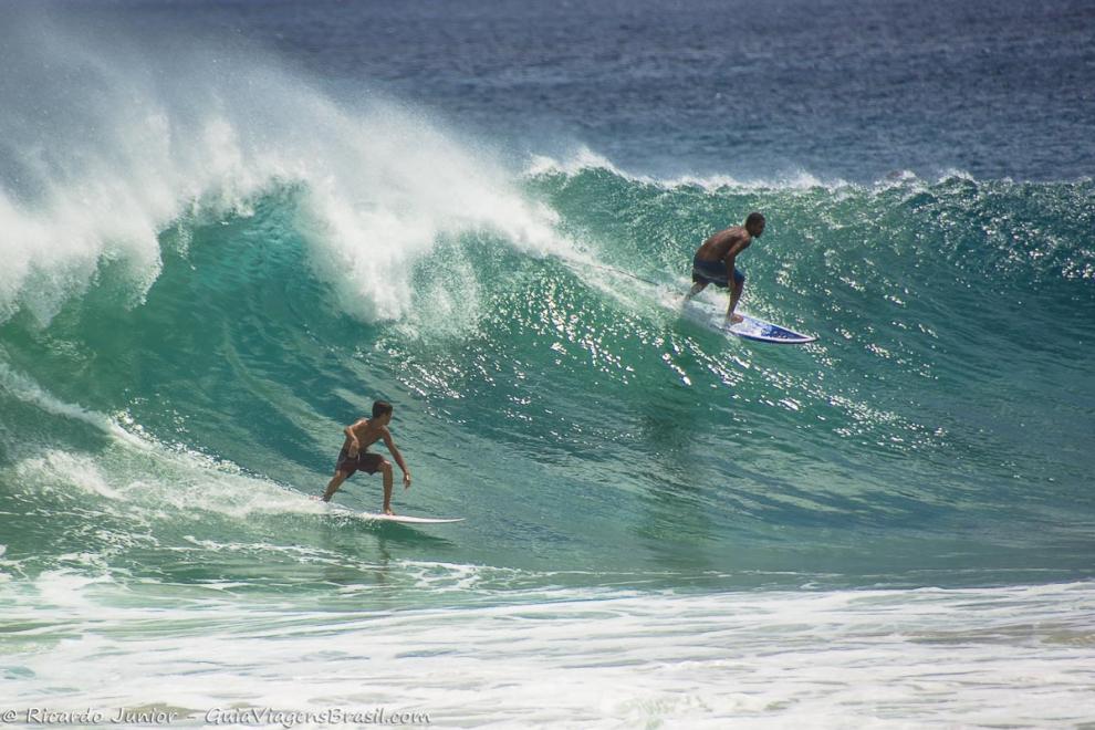 Imagem de dois surfista um no alto da onda e outro na base em Noronha.